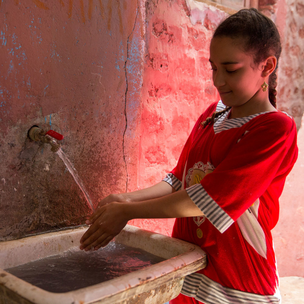 Esraa, 11, washes her hands in her home in Beni Edrees, while Heba talks to her mother about her satisfaction with the waste water services. She shared they now shower much more often as they no longer have to worry about their septic tank overflowing. Their tank used to fill up every week, which was very expensive to drain. Photo for USAID/Egypt.